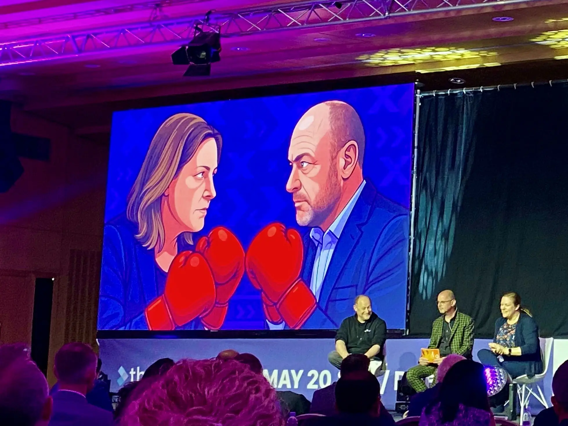 3 people sat on stage with a screen behind them with a cartoon of two people with red boxing gloves on